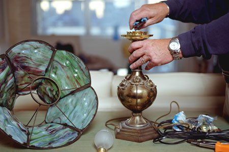 Hands repairing a brass lamp beside a large stained-glass lampshade and tools on a table