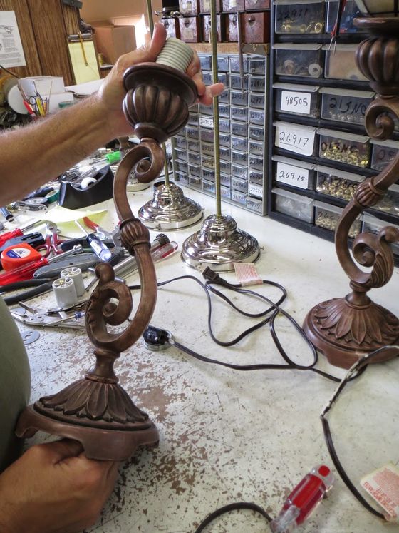 Hands holding a decorative bronze lamp base in a cluttered workshop with tools and shelves in the background