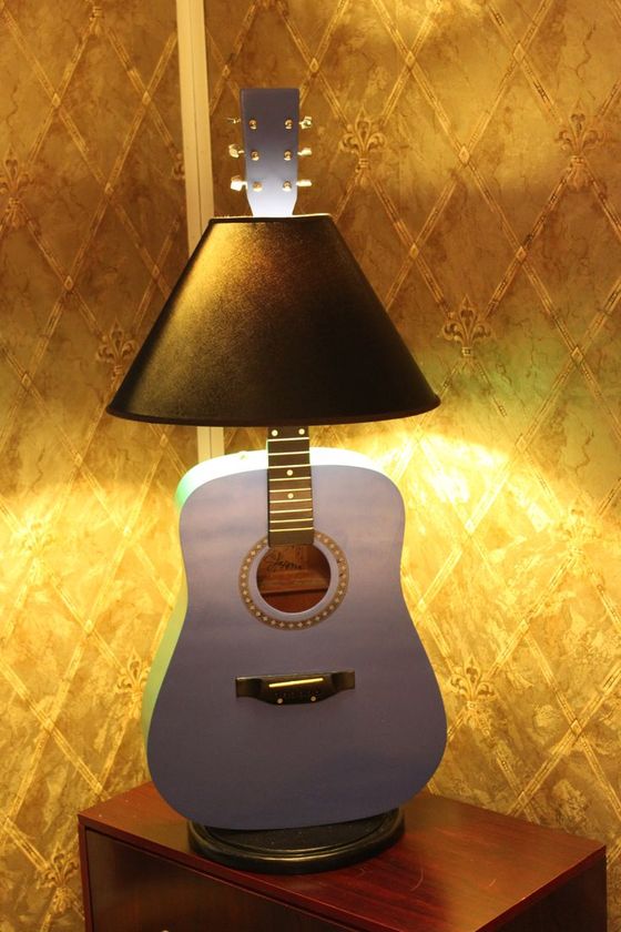 A guitar-shaped lamp with a black shade on a wooden table against a patterned wall.