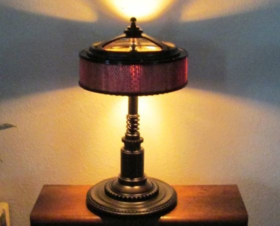 Lit table lamp with a red shade on a wooden surface, casting warm light and shadows on the wall.