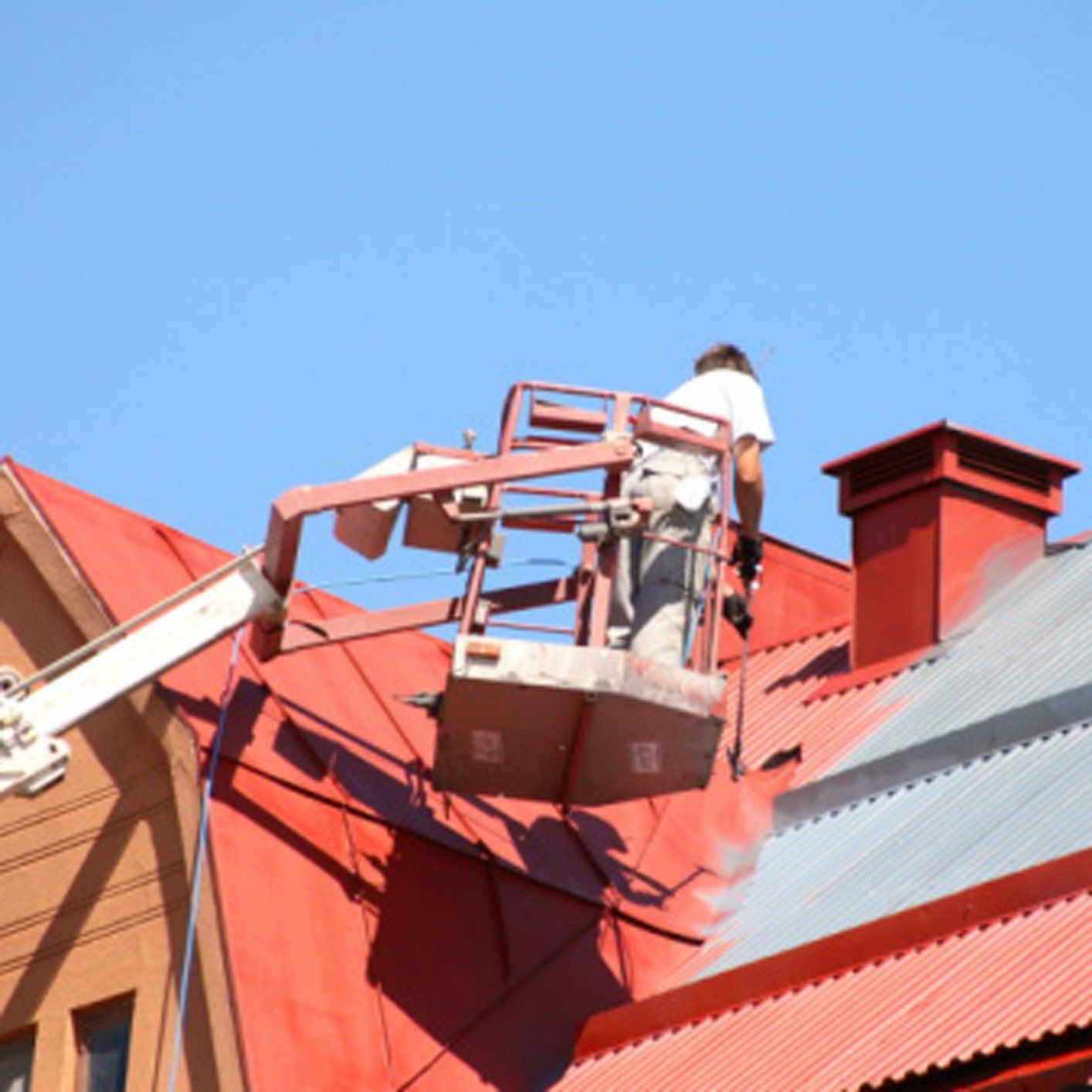 Worker in a lift repairing a red metal roof under a clear blue sky