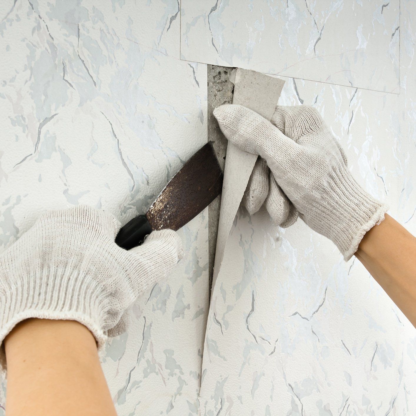 Gloved hands scraping old wallpaper from a wall with a putty knife