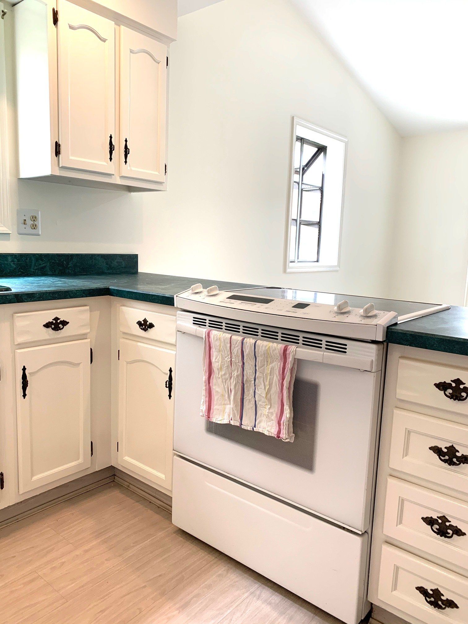 Bright white kitchen with cream cabinets, green countertops, and a stove beneath a window.