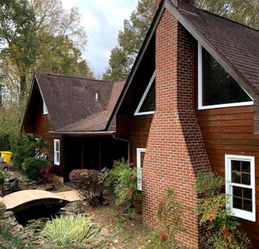 Two brown cabins with steep roofs and a brick chimney beside a small garden and footbridge