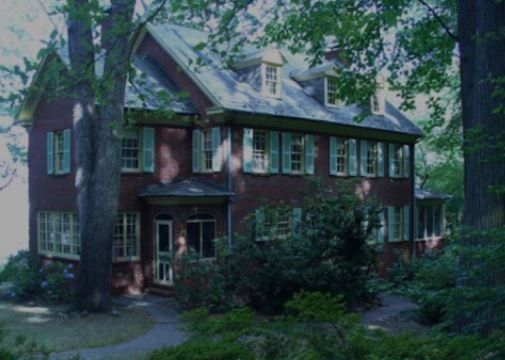 Two-story brick house with green trim, surrounded by trees and shrubs