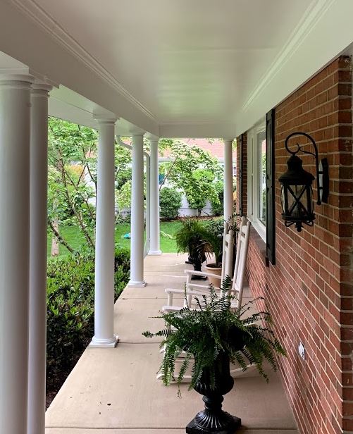 Covered front porch with white columns, brick wall, potted plants, and outdoor seating along a garden view.