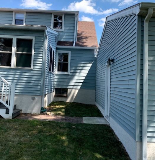 Narrow side yard between light blue vinyl-sided houses with grass and concrete walkway under a sunny sky