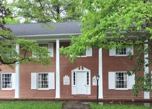 Brick colonial-style building with white columns, windows, and a central doorway under green trees