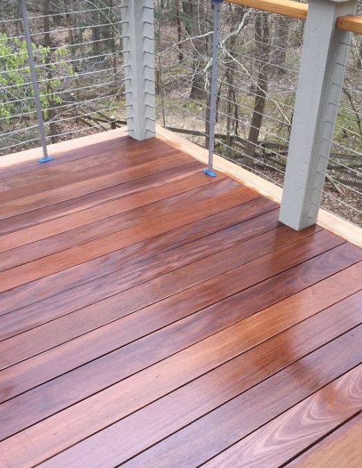 Wooden deck with reddish-brown planks, white posts, and wire railing overlooking trees outdoors