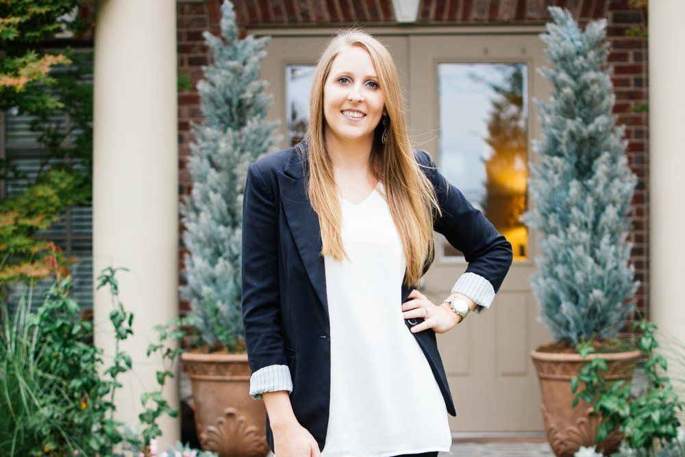 A woman in a black jacket and white shirt is standing in front of a brick building.