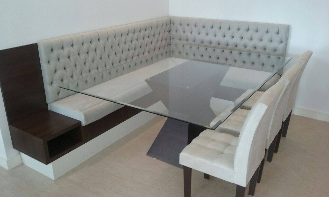 Mesa de canto para refeições com encosto estofado com botões, combinada com uma mesa de vidro e cadeiras.