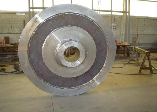 Un grande componente industriale metallico circolare con un foro centrale, appoggiato su un pavimento di cemento in un'officina.