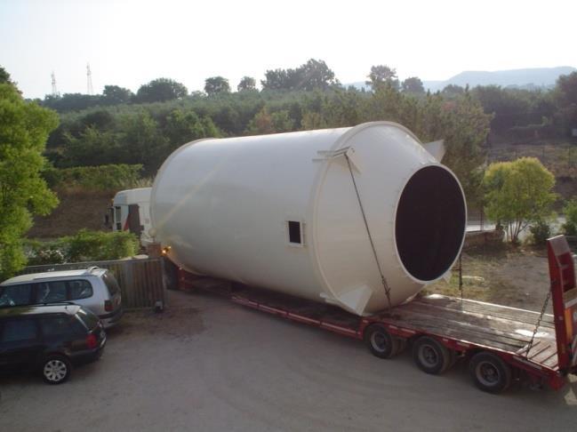 Un grande serbatoio industriale bianco viene caricato su un camion con pianale parcheggiato in un ambiente esterno alberato.
