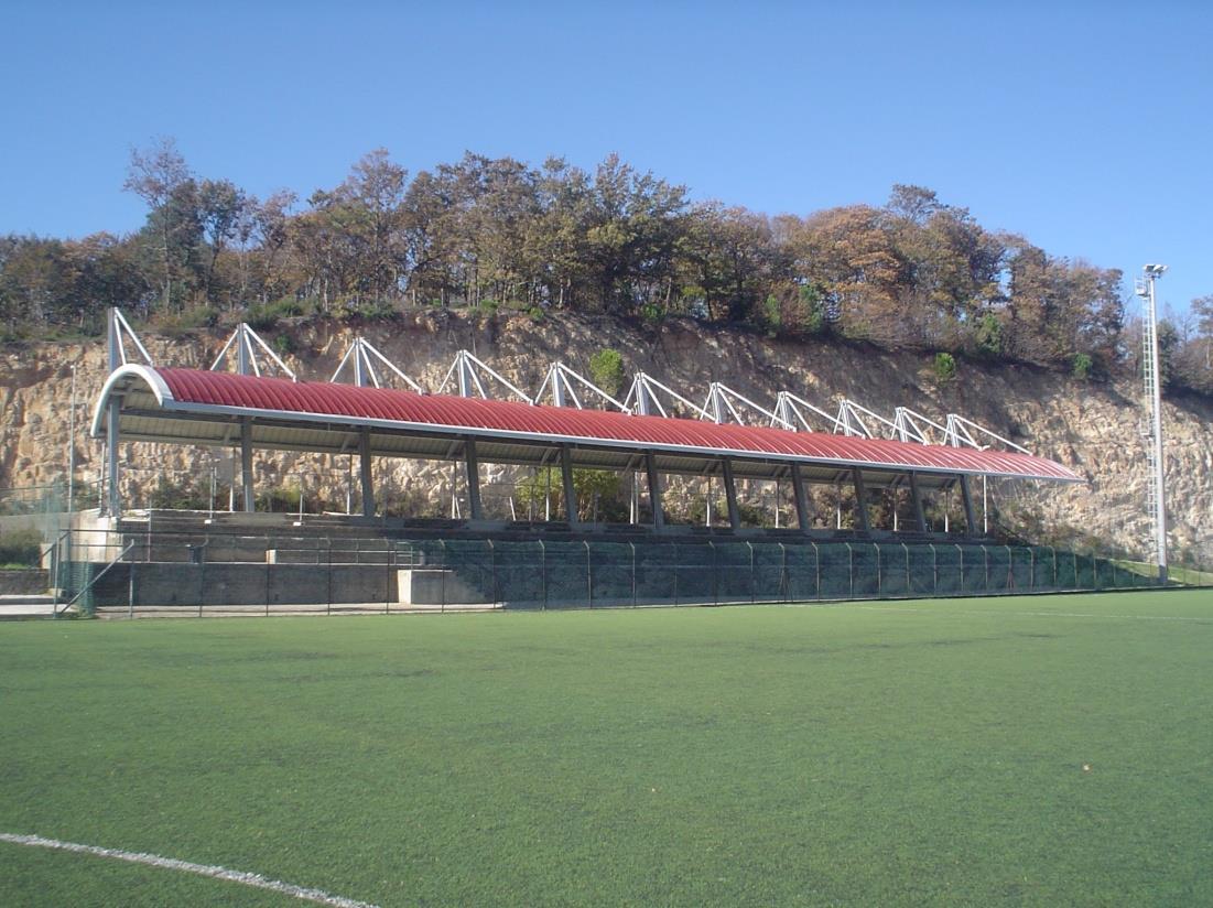 Un campo da calcio con una tribuna coperta dal tetto rosso, posizionato ai piedi di una collina erbosa e rocciosa.