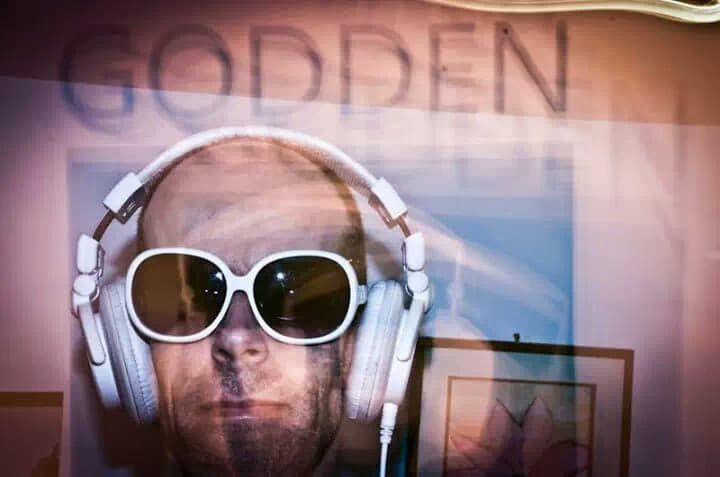 A man wearing headphones and sunglasses is standing in front of a sign that says godden.