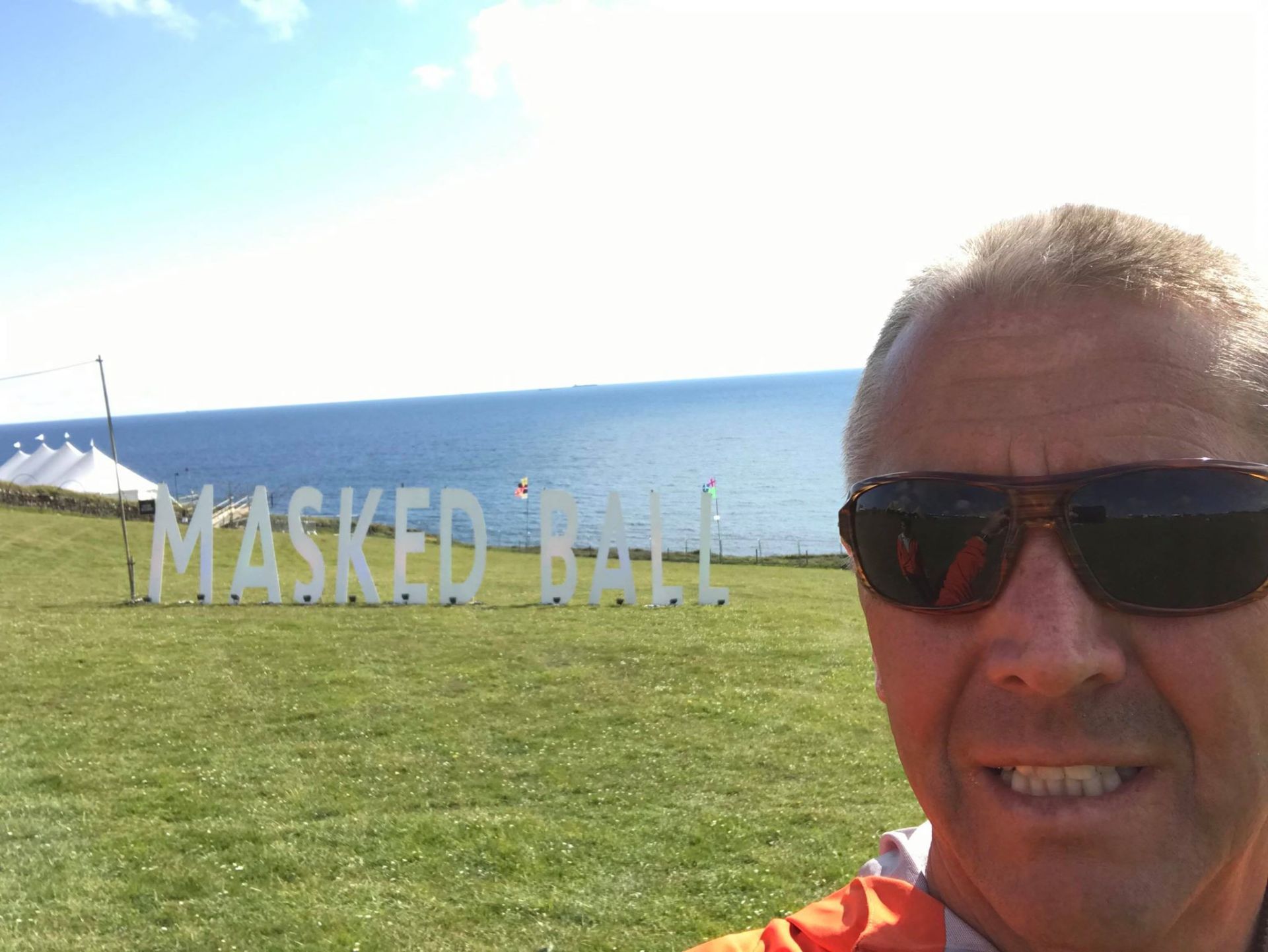 A man wearing sunglasses is standing in front of a sign that says masked ball.