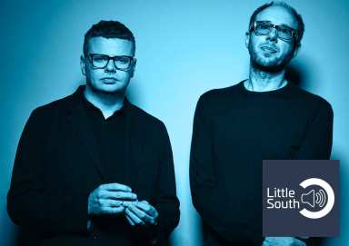 Two men wearing glasses are standing next to each other in front of a blue wall.