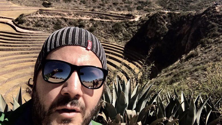 Um homem usando óculos escuros e um gorro está parado em frente a uma montanha.