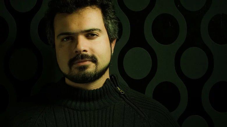 Um homem com barba está vestindo um suéter preto e olhando para a câmera.