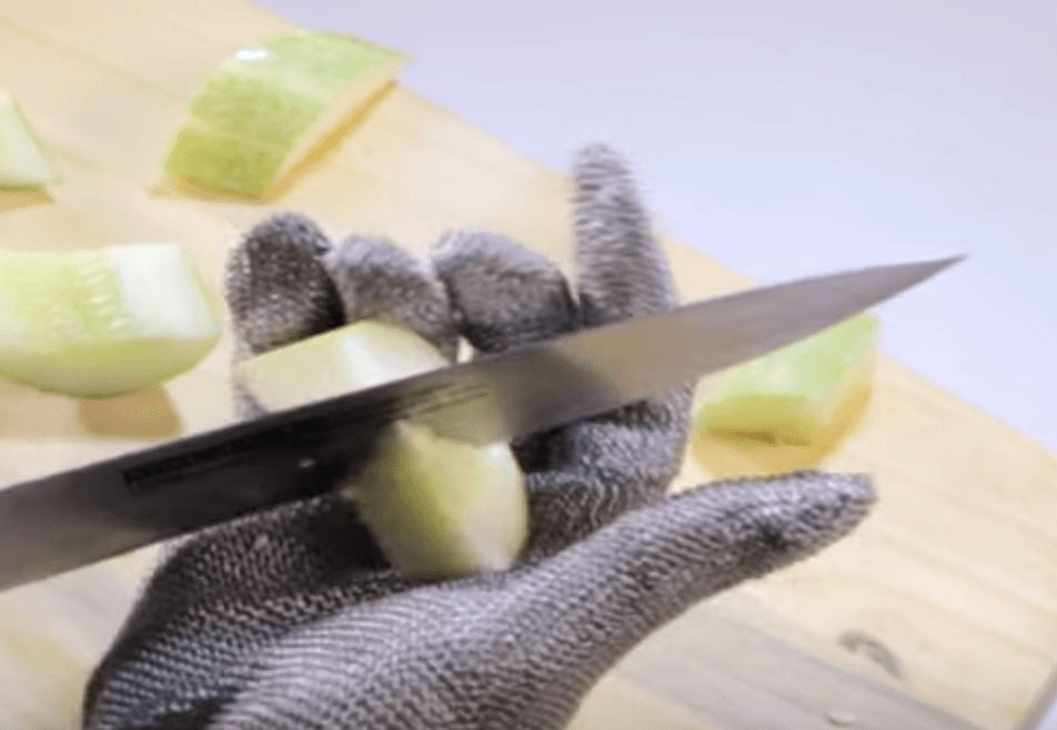 A person wearing a chain mail glove is cutting a cucumber on a cutting board.