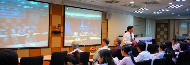 A man speaks to an audience in a conference room with two screens displaying an event.