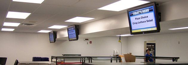 Three wall-mounted monitors display text in a cafeteria. Fluorescent lights illuminate the room.