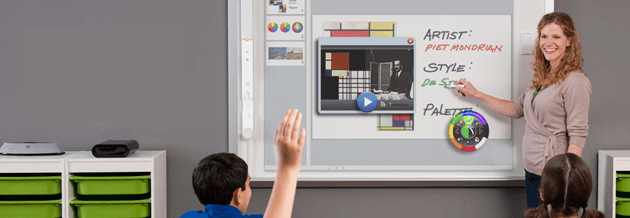 Teacher points to whiteboard, boy raises hand. Classroom setting with green storage bins.