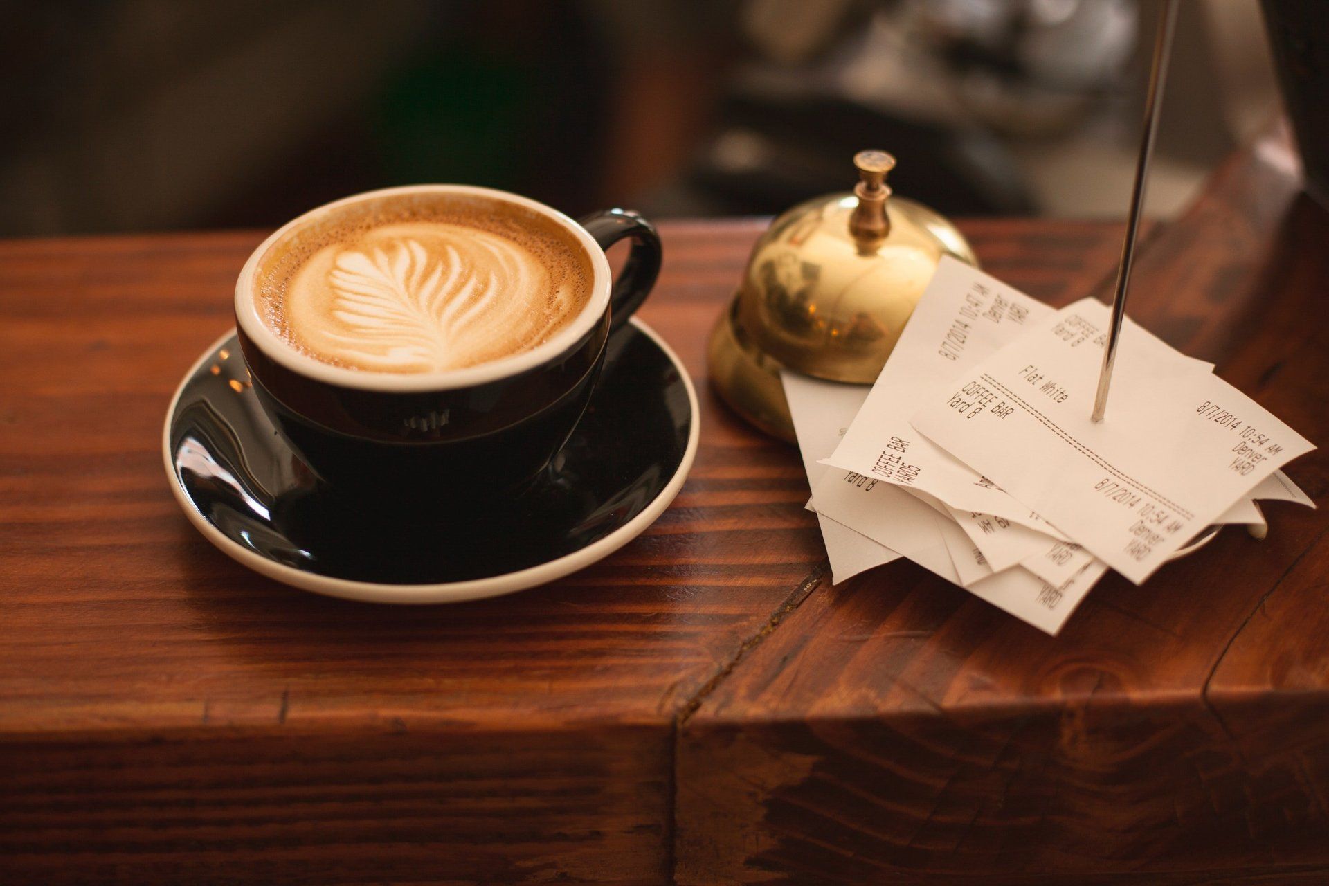 A cup of cappuccino is sitting on a wooden table next to a stack of receipts.
