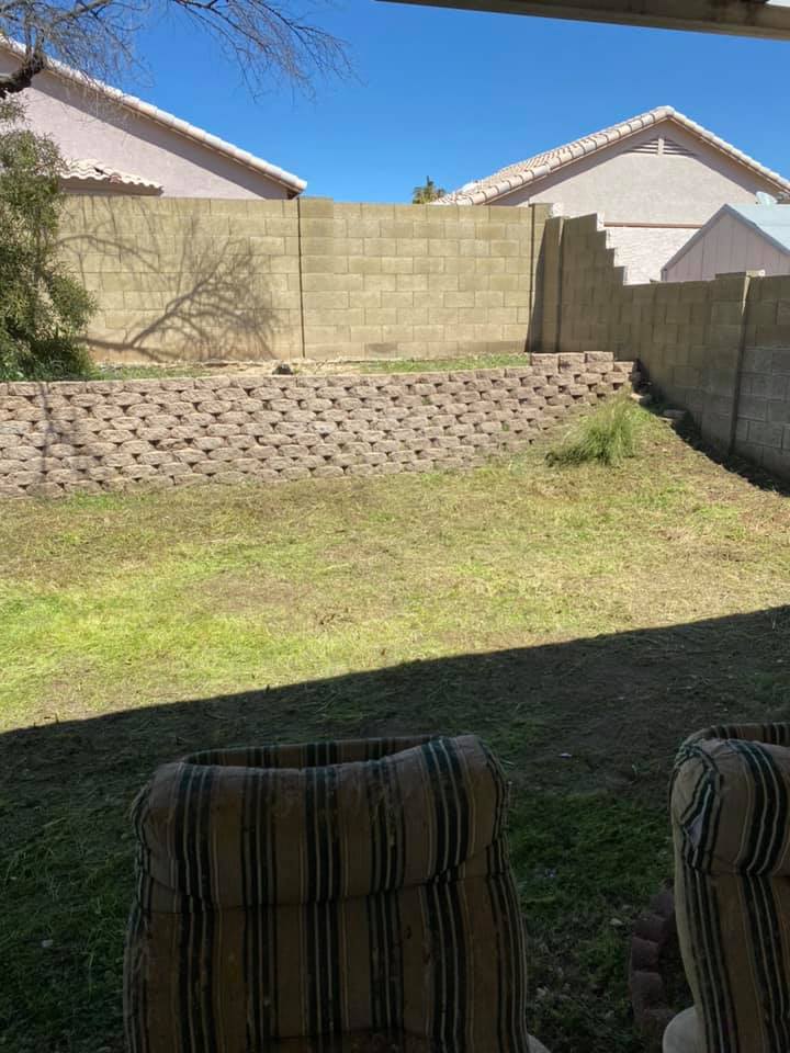 Backyard with grassy lawn, cinder block retaining wall, and beige privacy fence under bright sunlight