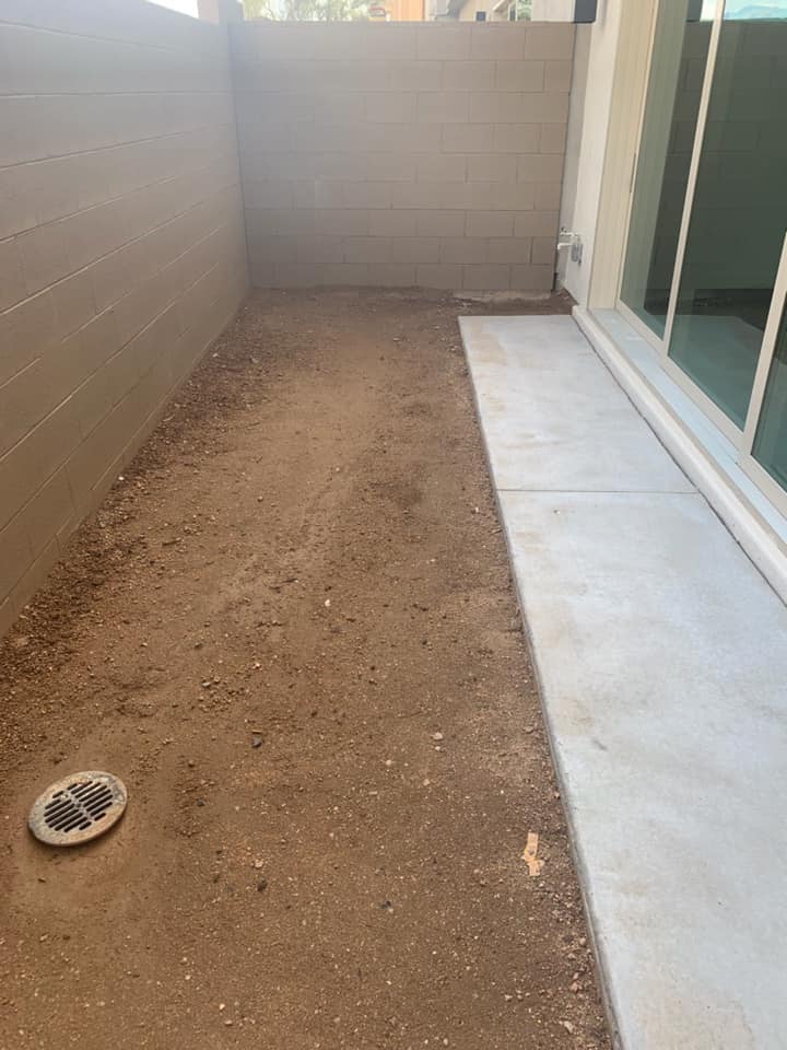 Narrow dirt side yard beside a concrete patio and sliding glass door, with block walls and a drain grate