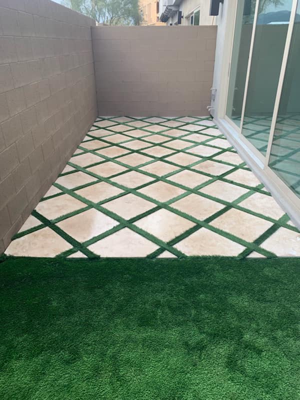 Small tiled patio with green diamond lattice, bordered by walls and glass doors, ending in artificial grass.