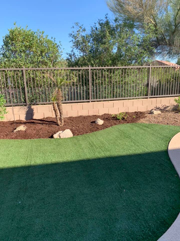 Backyard with green turf, tan retaining wall, metal fence, and trees under a blue sky