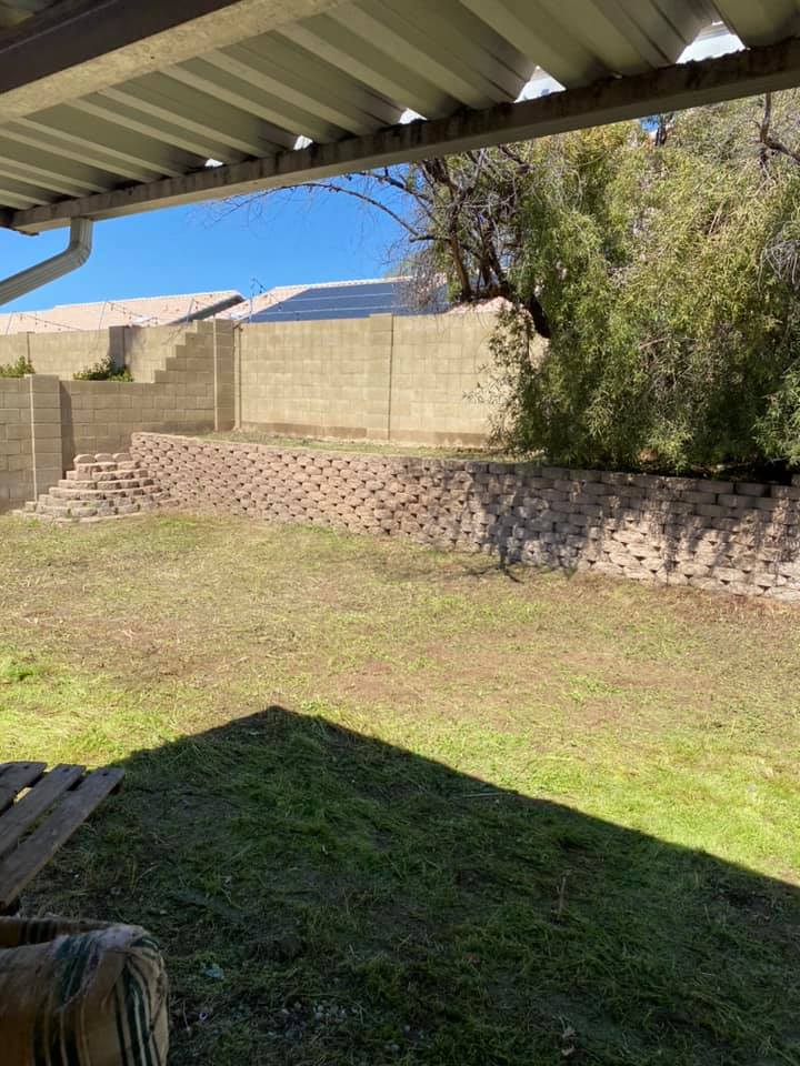 Backyard with green lawn, brick retaining wall, trees, and a covered patio roof casting shade