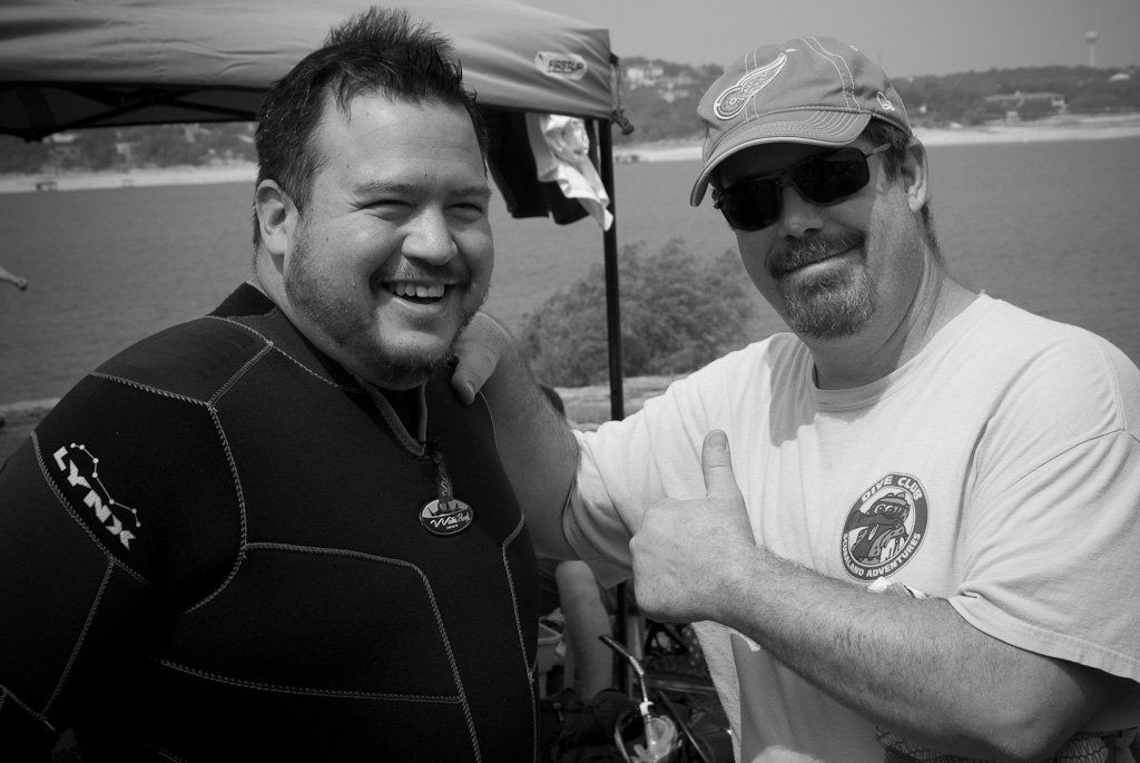 Two men smile near a body of water. One wears a wetsuit, the other a hat and sunglasses, giving a thumbs-up.