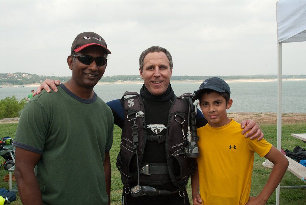Three people, two men and a boy, pose outdoors near a body of water. One man in scuba gear.