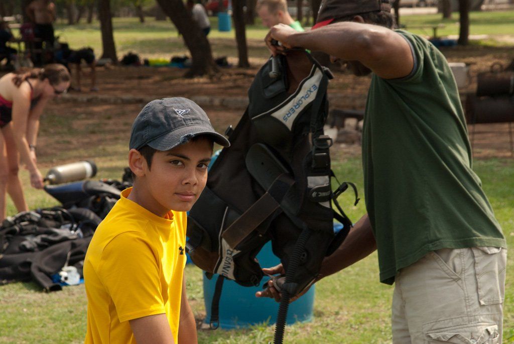 Boy in yellow shirt watches as man in green shirt adjusts scuba gear in a park.