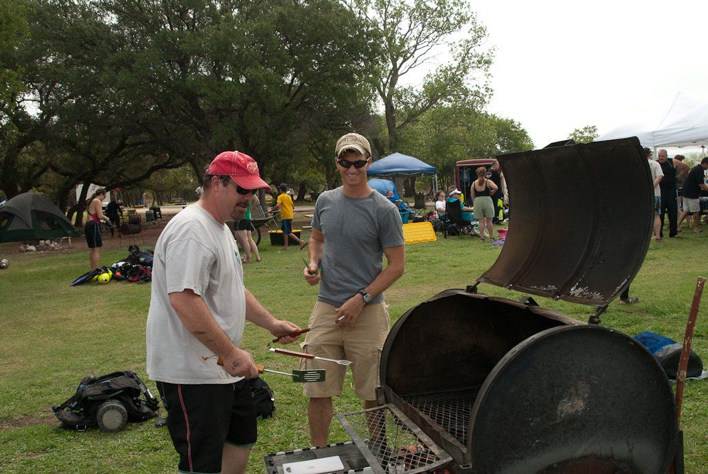 Two men grilling outdoors, one wearing a red cap, others in a park setting near tents and a canopy.