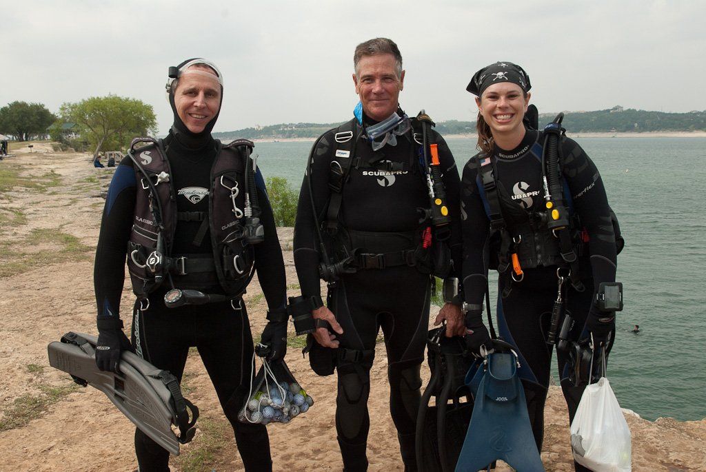 Three people in scuba gear stand near water, smiling.