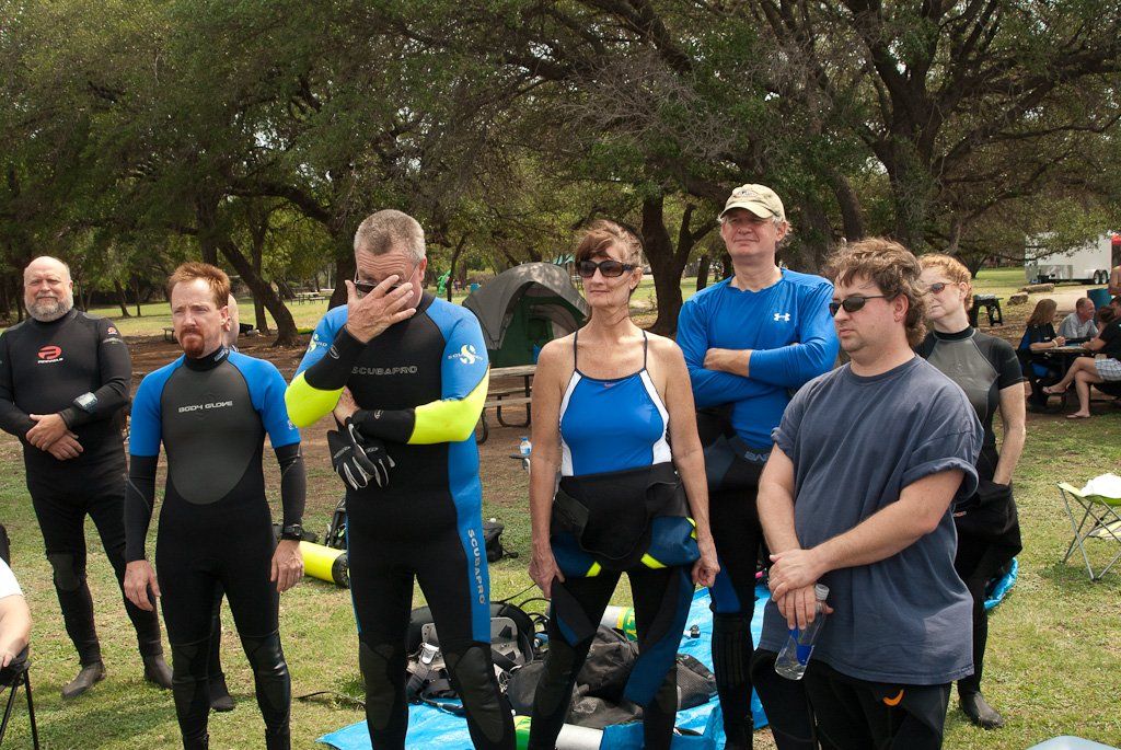 A group of people in wetsuits outdoors, preparing for diving. Some are smiling.