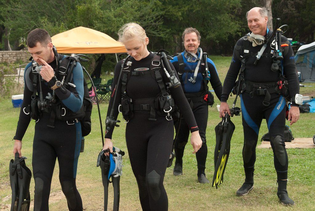 Four scuba divers in wetsuits walking on grass, carrying gear. An orange tent is in the background.