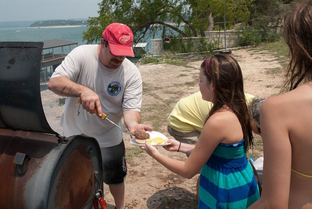 Man serving corn on the cob from a smoker to a woman in a blue dress, with a lake in the background.
