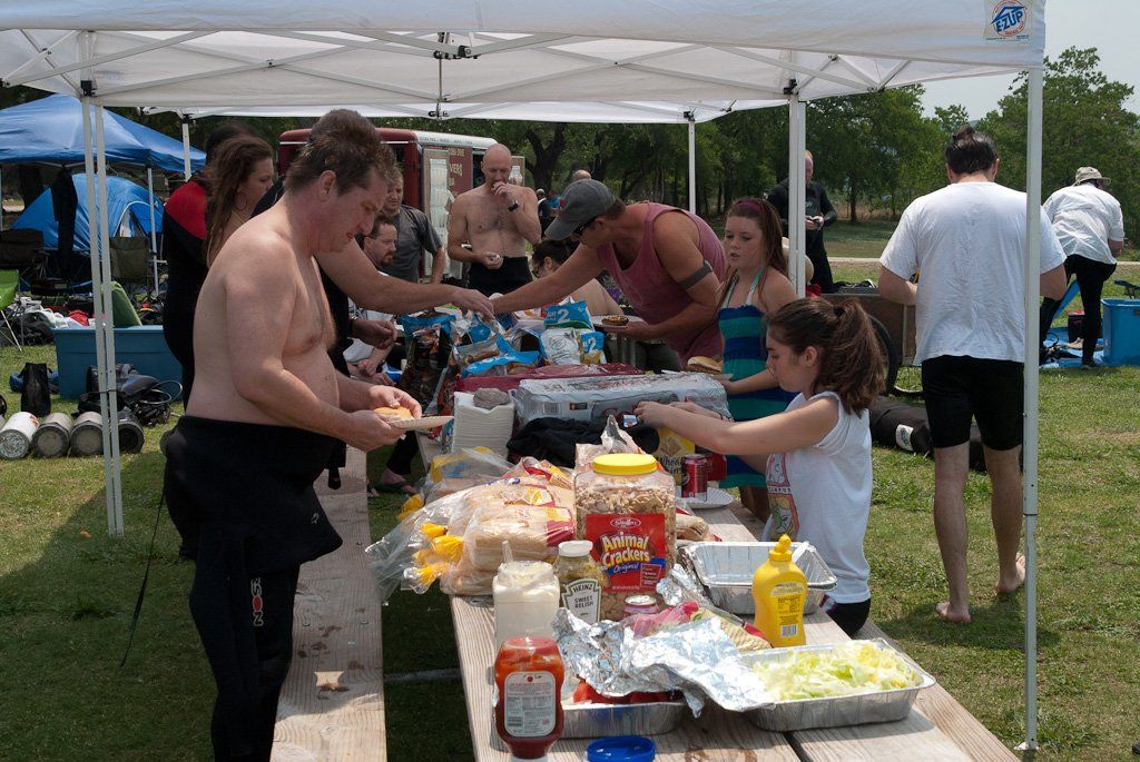 People at a picnic table under a canopy, preparing and serving food outdoors.