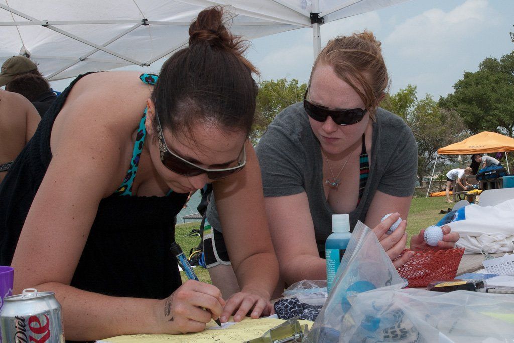 Two women wearing sunglasses, writing on paper at an outdoor event, one holds a pen, surrounded by items.