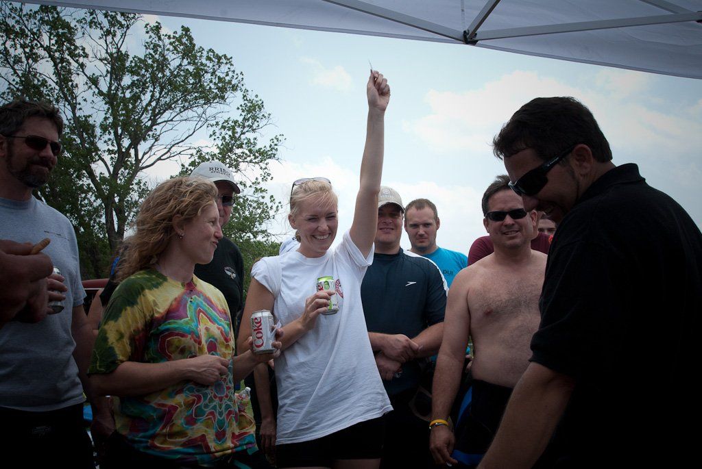 Group of people celebrating outdoors; woman with arm raised, holding a beverage, under a tent.