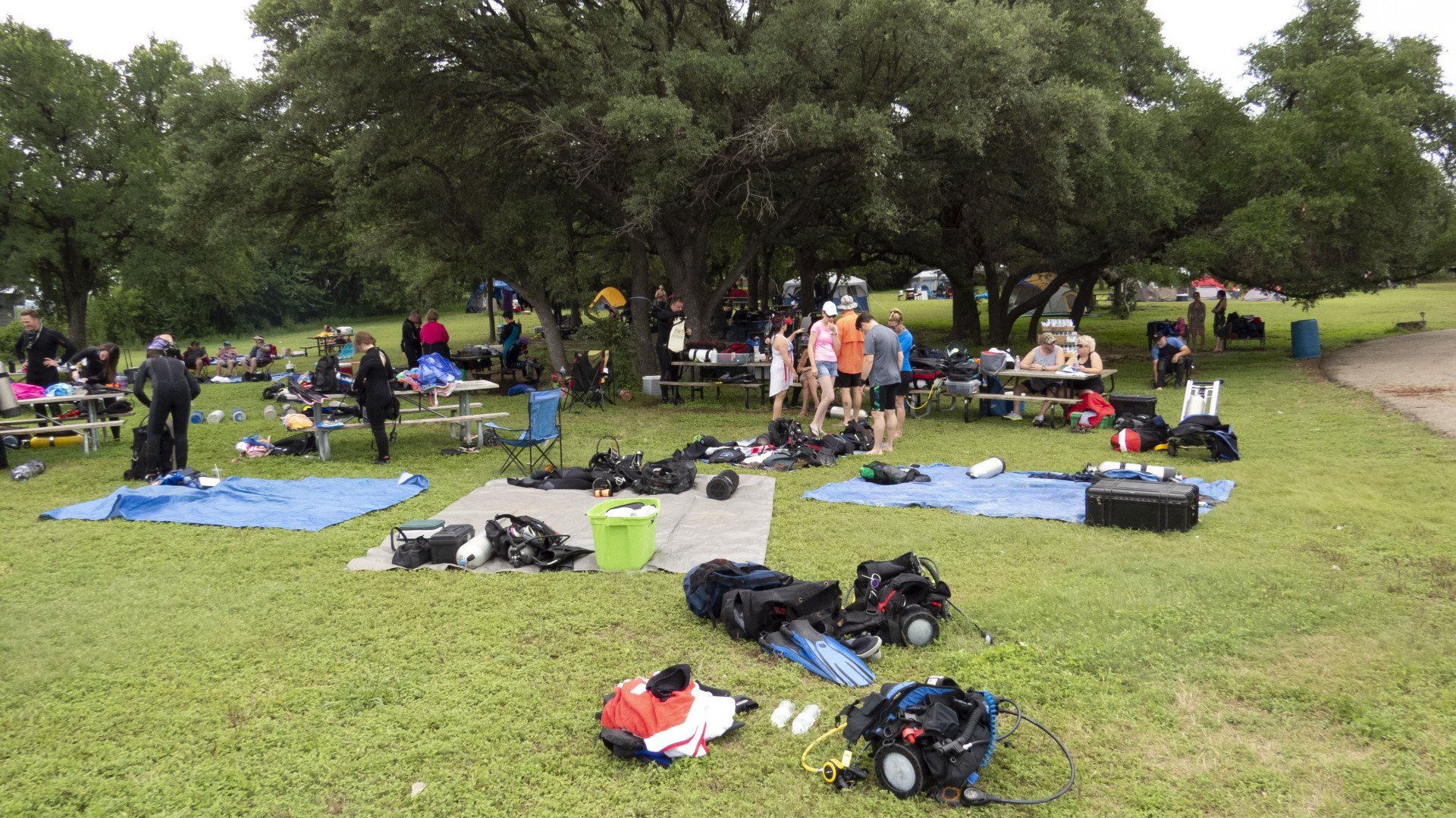 People gather outdoors near trees with equipment laid out on the grass and tables.