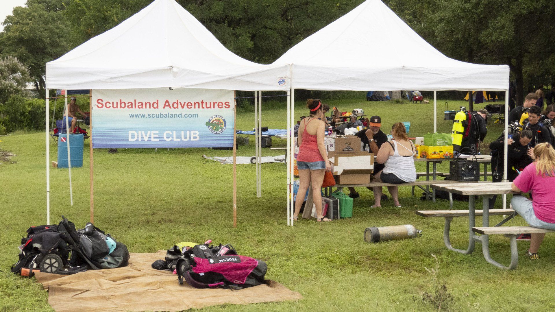 Two white tents with people gathered, picnic table, gear on the ground, sign reads 