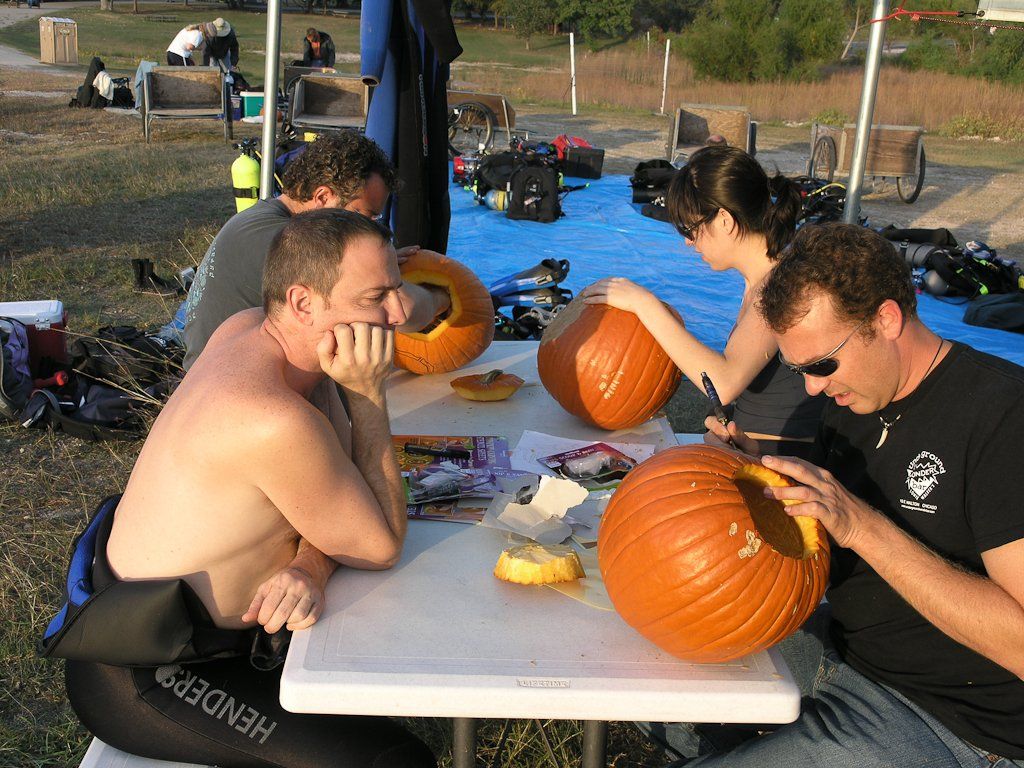 People carving pumpkins outdoors. A man in a wetsuit looks on while others carve pumpkins on a table.