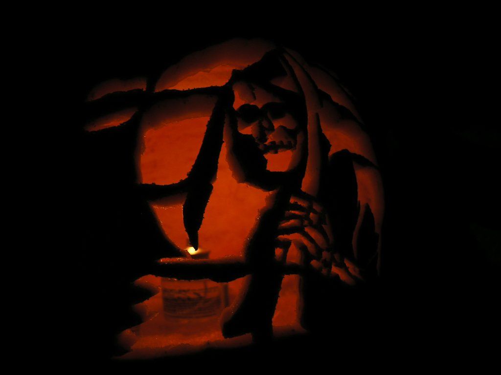 Jack-o'-lantern carved with a grim reaper design. Lit from within, showing skull, scythe, and a cloaked figure.