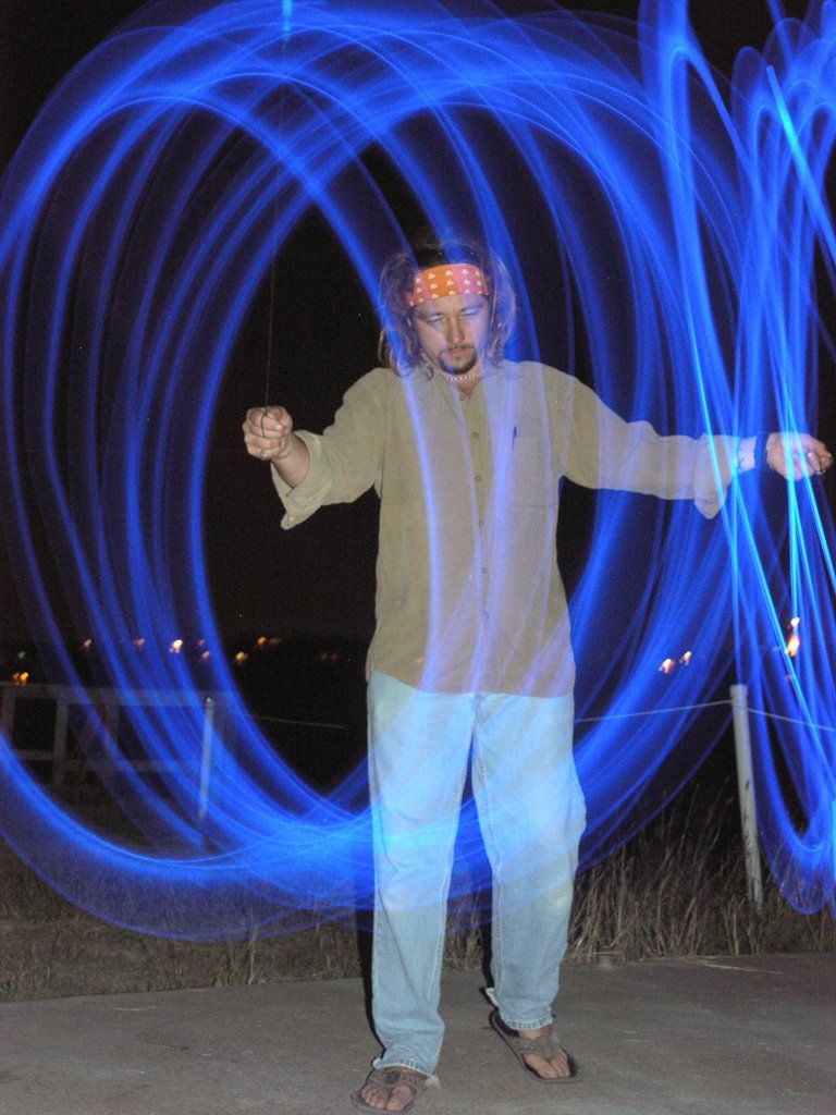 Man with long hair and headband spins a glowing blue light in circles at night.