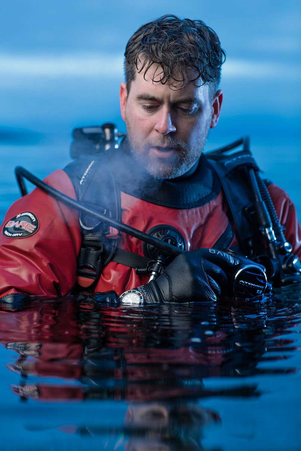 Scuba diver in red drysuit emerges from blue water, holding equipment, looking focused.